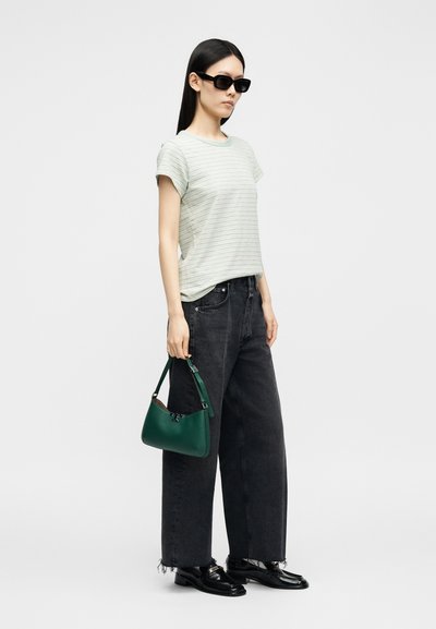 Vrouw met een zwarte zonnebril, lichtgrijs gestreept t-shirt, losse zwarte jeans, glanzende zwarte schoenen, een donkergroene handtas naast zich houdend.