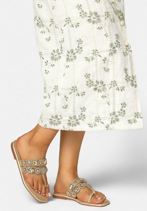 Pies con sandalias planas beige con piedras decorativas y tiras trenzadas, combinadas con una falda blanca bordada con motivos florales.