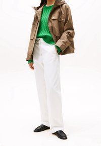 Bruine capuchonjas met twee zakken aan de voorkant, groene geribde trui, witte jeans met wijde pijpen en zwarte platte schoenen.