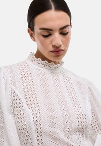 Blouse blanche avec broderie ajourée complexe, col montant à bord festonné et manches bouffantes, confectionnée dans un tissu léger.