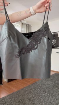 Svart satin camisole med spetsapplikation, hålls av tatuerade armar som bär en vit smartklocka, i ett modernt vardagsrum.