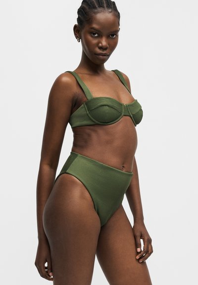 Femme aux cheveux tressés portant un bikini côtelé vert olive avec un haut à armatures et un bas taille haute, debout devant un fond uni.