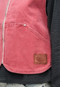 Κόκκινο υφασμάτινο γιλέκο με διαγωνιακή τσέπη και μεταλλική λεπτομέρεια κουμπιού, φορεμένο πάνω από ένα σκουρόχρωμο πουκάμισο με σχέδια, με καφέ ταμπελάκι που γράφει «element since 1992».