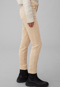 Pantaloni corduroy beige con vestibilità slim, dotati di tasche frontali e zip. Abbinati a stivali neri alla caviglia con superficie texturizzata.