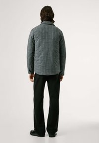 Una persona de pie de espaldas al espectador, vistiendo una chaqueta a cuadros, pantalones negros y zapatos, contra un fondo claro y neutro.