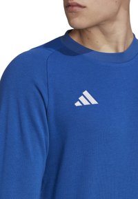 Blauwe sweater van zacht materiaal, met een ronde halslijn, ribbelkraag en een wit Adidas-logo aan de bovenlinke kant.