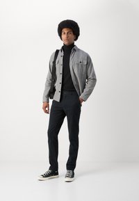 Chemise en flanelle grise par-dessus un col roulé noir, associée à un pantalon sombre et des baskets noires. Présente une poche poitrine et un design à boutons.