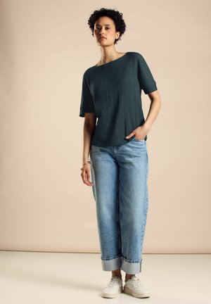 Dunkelgrünes, strukturiertes Kurzarmoberteil kombiniert mit hellblauen, locker sitzenden Jeans und weißen Sneakern, accessoriert mit einem Armband.