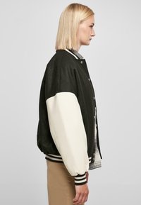 Čierny a biely varsity kabát s patentným zapínaním, kontrastnými rukávmi, rebrovanými manžetami a jemnou textúrou, nosený cez károvanú košeľu.