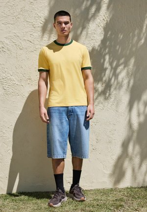 Junger Mann in gelbem T-Shirt und blauen Jeansshorts steht vor einer strukturierten beigen Wand mit Schatten auf dem Gras.