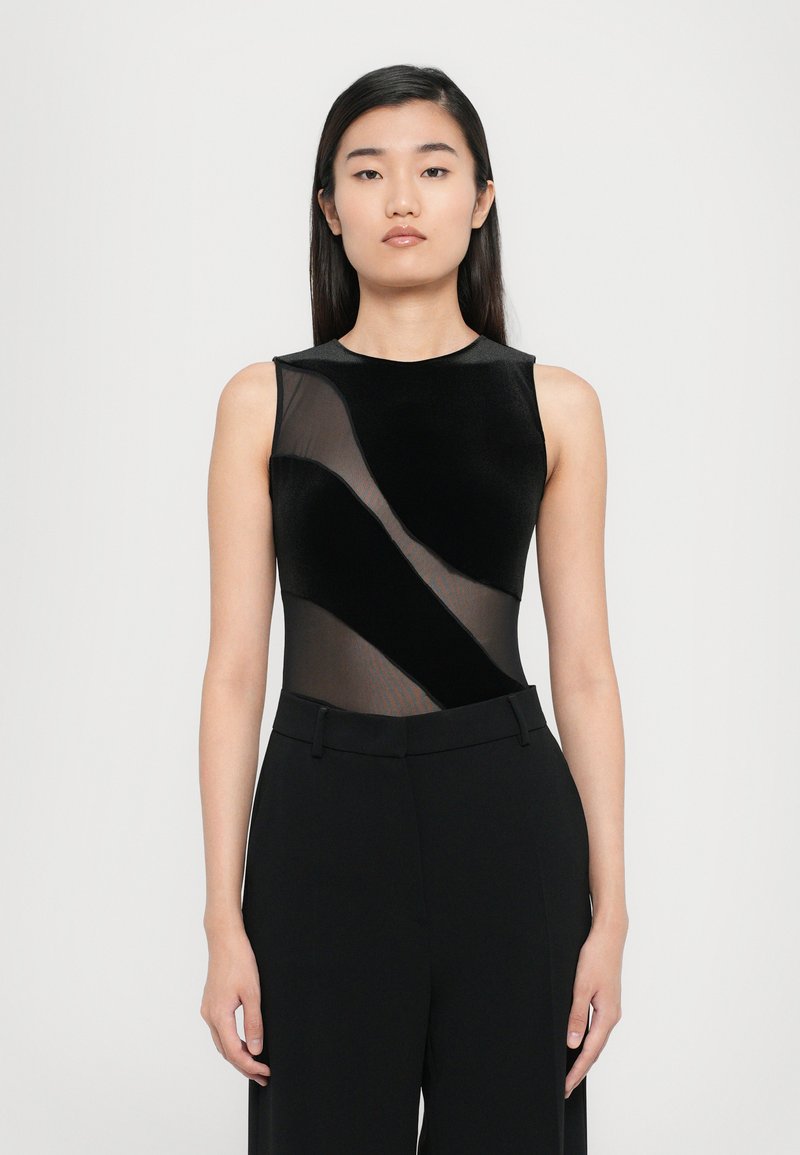 Black bodysuit with a sheer mesh panel design. Features a sleeveless cut, rounded neckline, and smooth texture, paired with high-waisted black trousers.