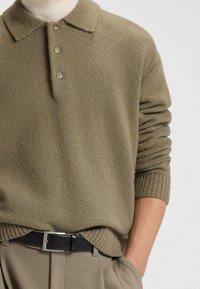 Polo in maglia verde oliva con finitura texturizzata, tre bottoni e polsini a coste, abbinato a pantaloni beige su misura e una cintura nera.