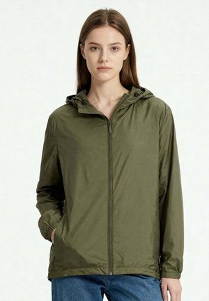 Jonge vrouw met steil bruin haar, gekleed in een olijfgroene zip-up capuchonjas en blauwe jeans, staand met één hand in de zak.