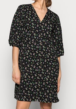 Femme portant une robe noire à encolure en V avec un petit imprimé floral violet, vert et beige, manches trois-quarts bouffantes, se tenant devant un fond blanc.