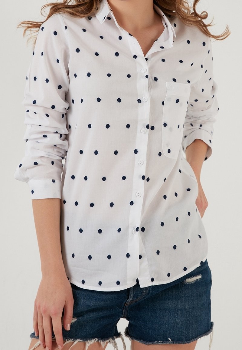 Femme portant une chemise blanche à boutons avec des pois noirs et des manches retroussées, associée à un short en denim effiloché.