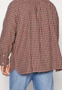 Camisa de cuadros en tonos rojos, blancos y marrones, con un corte relajado, puños abotonados y una abertura en la parte posterior para mayor libertad de movimiento.