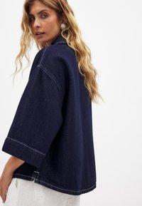 Kvinde med langt blont hår iført sølvspiraløreringe, en løs mørkeblå denimjakke og hvide bukser, som ser over sin skulder.