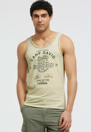 Mann trägt ärmelloses beiges Tanktop mit dem Text "Camp David" und grüne Cargo-Shorts, steht mit einer Hand in der Tasche vor weißem Hintergrund.