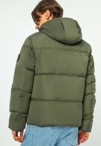Oliwkowa kurtka puchowa z kapturem, z pikowanym wzorem i okrągłą czarną łatą z logo na lewym ramieniu. Miękka tekstura, luźny krój.