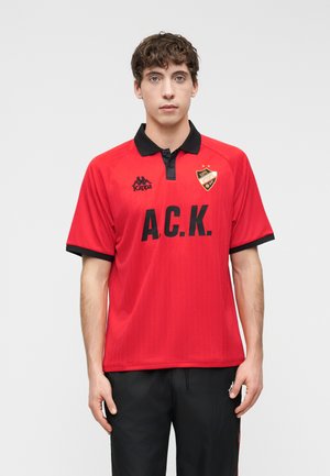 Mladi moški, oblečen v rdečo Kappa polo majico s črnim ovratnikom, napisom "A.C.K.", logotipi in črnimi hlačami na nežni svetli podlagi.