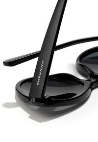 Black sunglasses with an oval-shaped frame and dark lenses. Made of plastic, features slim black temple arms, with "HAWKERS" logo on one arm.