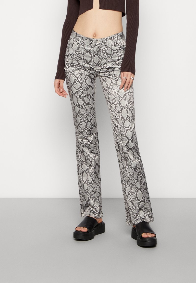 Spodnie flare z nadrukiem w wężową skórę w kolorze srebrnym i czarnym, charakteryzujące się gładką teksturą i dopasowanym pasem. Noszone z czarnymi sandałami na platformie.