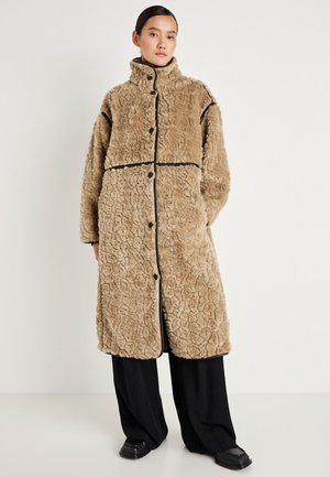 Bežinis dirbtinio kailio paltas su tekstūruotu dizainu, aukštu apkakle, juodais akcentais ir sagomis priekyje, derinamas su plačiomis juodomis kelnėmis.