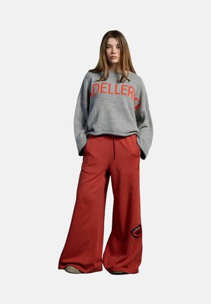 Jeune femme debout, les mains dans les poches, portant un pull gris avec un texte rouge et un pantalon large rouge avec un patch près du genou.