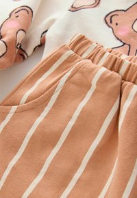 Läheltä kuvattua pehmeää kangasta, jossa on beigellä ja valkoisella pystysuoria raitoja sekä kermaista puuvillavaatetta, jossa on piirroshahmo karhusta.