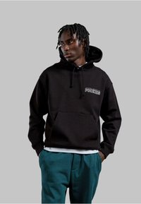 Felpa nera in cotone con tasca frontale e cappuccio con cordino, caratterizzata da testo bianco sul petto. Abbinata a pantaloni color teal.