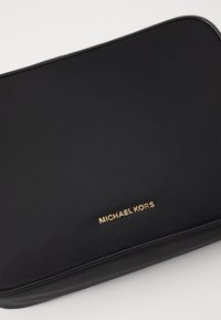 Melnā sintētiskā soma ar gludu tekstūru, kurai apakšā ir zelta "MICHAEL KORS" logo, taisnstūra forma un noapaļoti stūri.