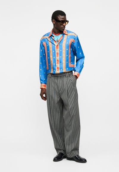 Bărbat purtând o cămașă albastră oversized cu model, cu accente aurii și roșii, pantaloni gri largi cu dungi fine, pantofi negri și ochelari de soare întunecați, stând în picioare.