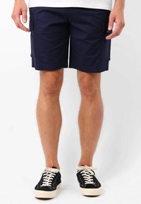 Shorts cargo bleu marine avec poches latérales, fabriqués en tissu lisse, dotés d'une coupe droite et d'ourlets cousus. Associés à des baskets noires.