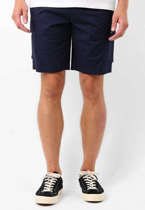Shorts cargo bleu marine avec poches latérales, fabriqués en tissu lisse, dotés d'une coupe droite et d'ourlets cousus. Associés à des baskets noires.