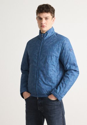 Modra zip-up jakna z teksturiranim vzorcem, visokim ovratnikom in logotipom, v kombinaciji s temnimi jeans hlačami. Mehka, lahka tkanina.