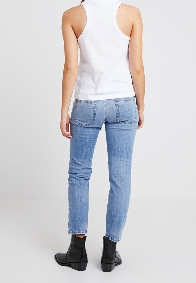 Femme portant un haut blanc sans manches, un jean bleu clair et des bottines noires, debout de dos sur un fond uni.