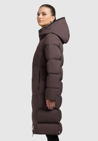 Brun pufferjakke med hætte, der har et langt design, kviltet tekstur og sidelommer. Indersiden af stoffet er sort.