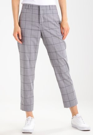 Unterkörper einer Person, die graue kariert zulaufende Hosen und weiße Turnschuhe trägt, stehend vor einem einfarbigen weißen Hintergrund.