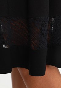 El dobladillo de un vestido negro con un panel de encaje negro transparente, que se lleva por encima de la rodilla, mostrando parte de la pierna de una persona.