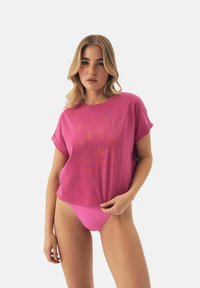 Różowa koszulka crop z podwiniętymi rękawami, z graficznym nadrukiem palm. W parze z różowymi, prążkowanymi spodniami typu high-waist. Gładka tekstura.