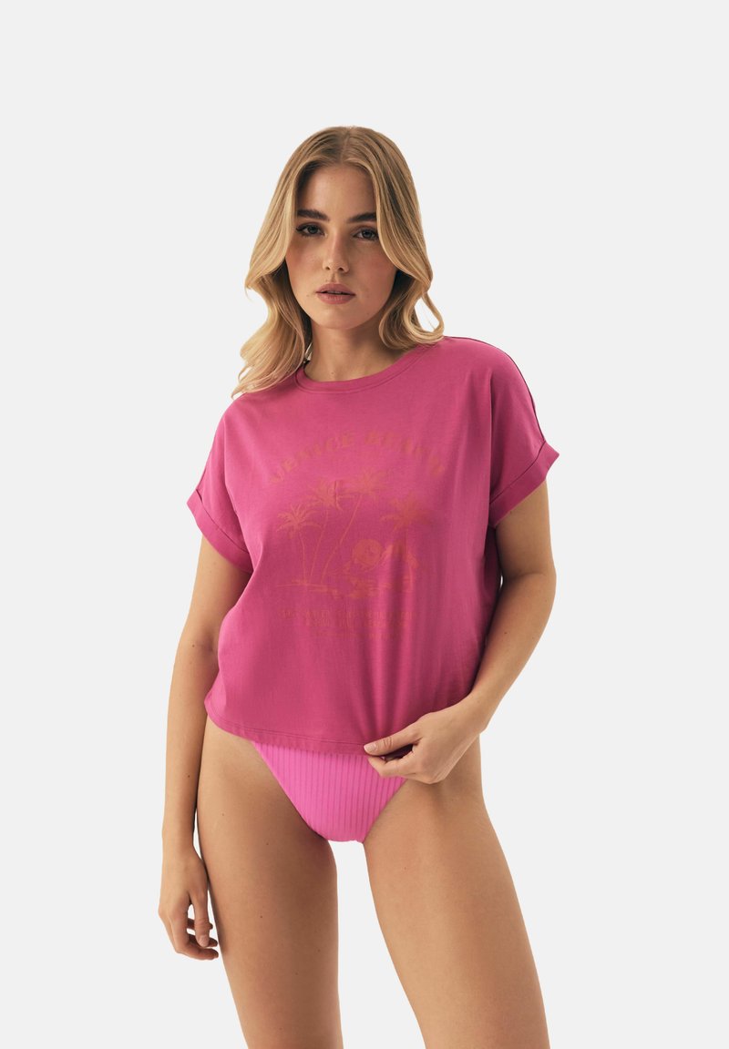 Różowa koszulka crop z podwiniętymi rękawami, z graficznym nadrukiem palm. W parze z różowymi, prążkowanymi spodniami typu high-waist. Gładka tekstura.