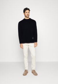 Maglione nero a maglia con scollo rotondo e maniche lunghe, abbinato a chinos crema e scarpe scamosciate marrone chiaro. Presenta un'etichetta con logo.
