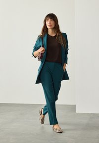 Traje de color verde azulado con blazer entallado y pantalones a medida, combinado con una camiseta marrón y zapatos planos con estampado. La modelo lleva un bolso pequeño y oscuro.