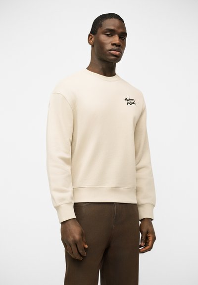 Άνδρας που φοράει μπεζ φούτερ με το λογότυπο "Maison Kitsuné" στο στήθος και καφέ παντελόνι, στέκεται μπροστά σε απλό φωτεινό φόντο.