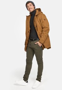Brun isolert jakke med hette, svart genser med høy hals, olivengrønne bukser og hvite joggesko med svarte detaljer. Modell står med avslappet holdning.