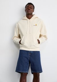 Krémová bavlnená mikina s kapucňou a šnúrkami, so prednou kapsou a malým logom Carhartt, v kombinácii s tmavými džínsoými šortkami.