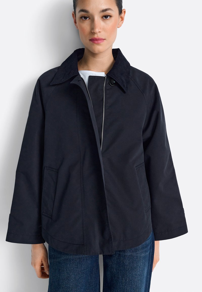 Tamsiai mėlyna striukė su tekstūruotu apykakliu, uždarymu priekyje su sagomis ir šoninėmis kišenėmis. Turi ilgus rankogalius ir laisvo kirpimo dizainą.