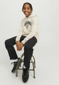 Bež hoodie s logotipom "JACK & JONES", crne hlače i crne tenisice; sjedi na metalnoj stolici nasuprot jednostavne pozadine.