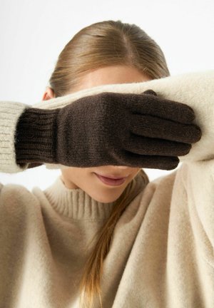 Brown and cream knit gloves cover part of a person's face. Gloves have ribbed cuffs and feature a textured pattern. Sweater is beige.