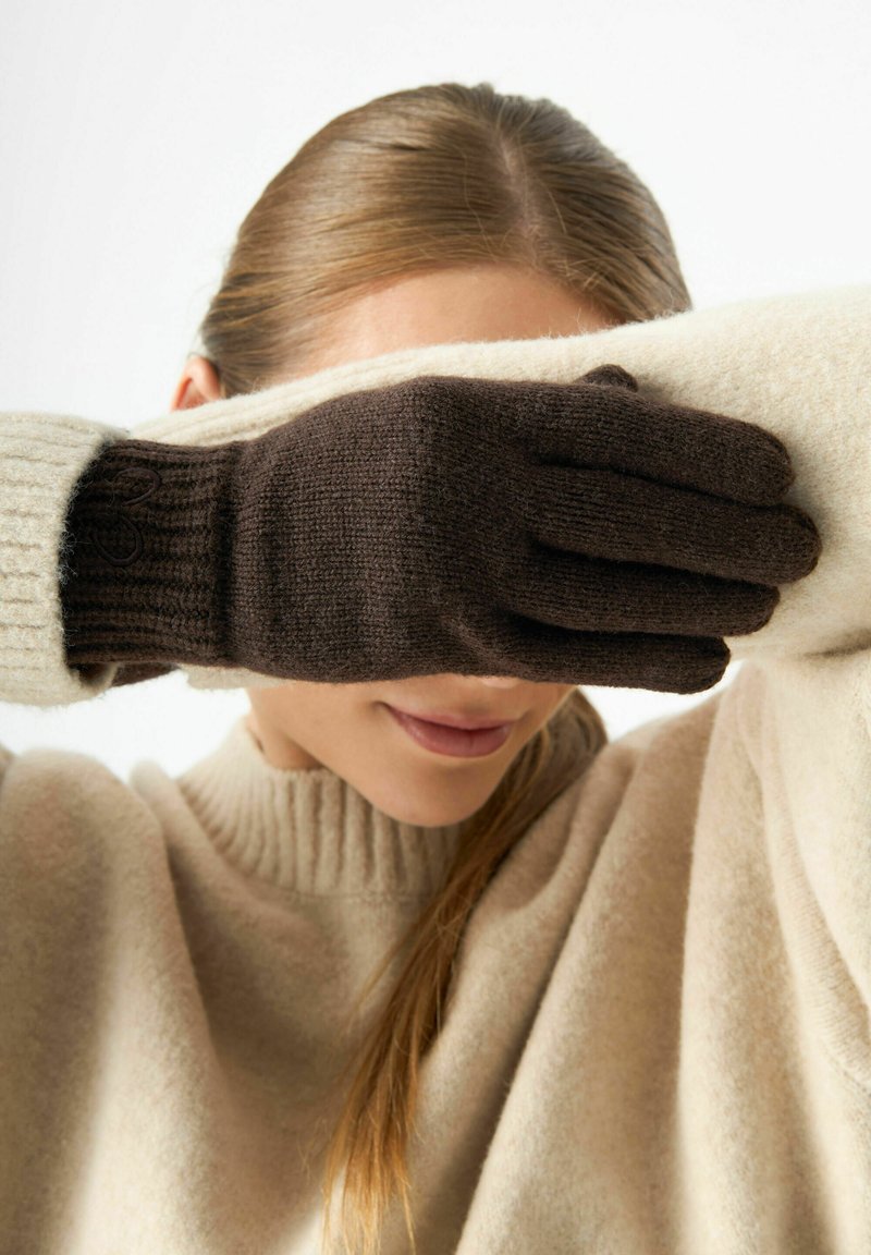 Braune und cremefarbene Strickhandschuhe bedecken einen Teil des Gesichts einer Person. Die Handschuhe haben gerippte Bündchen und weisen ein strukturiertes Muster auf. Der Pullover ist beige.